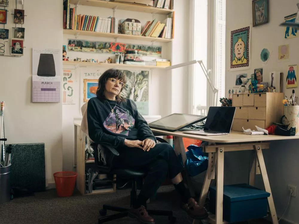Une personne est assise dans un bureau ou un atelier, entourée de livres, d'affiches et de divers objets. Elle porte des lunettes et un pull à motif. Sur le bureau, il y a un ordinateur portable et une tablette graphique.