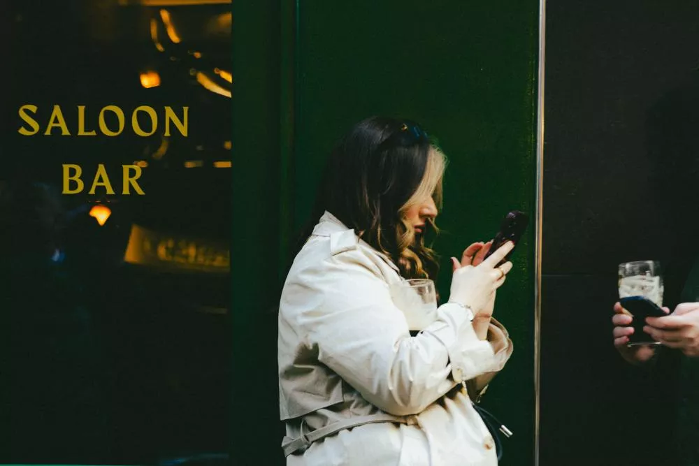 Une femme en manteau beige est debout devant un bar, absorbée par son téléphone. Elle tient un verre à moitié plein, tandis qu'une autre personne à côté d'elle tient également un verre et un téléphone.