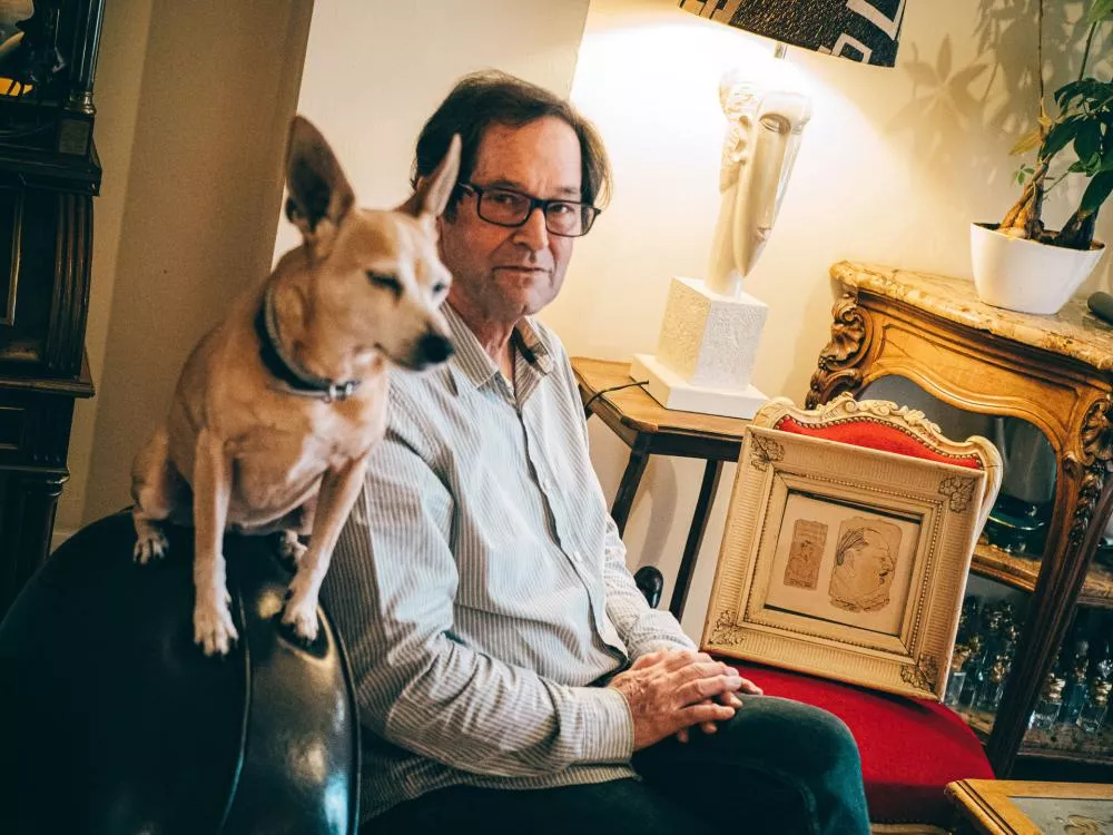 Un homme assis dans une pièce décorée, avec un chien à ses côtés. Il y a des meubles anciens, une lampe design et un cadre avec des dessins.