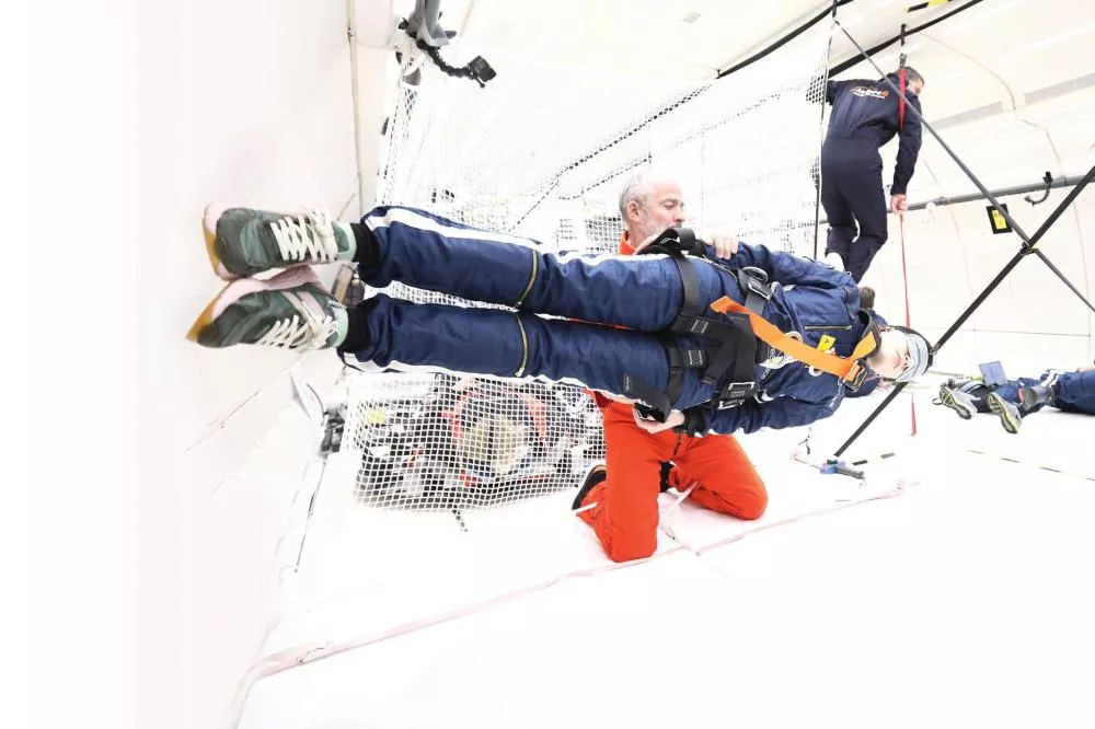 Un individu en combinaison bleue est suspendu horizontalement, soutenu par un harnais et accompagné d'une personne en combinaison orange à genoux. D'autres personnes en arrière-plan semblent participer à une activité similaire dans un environnement équipé de filets et de structures de soutien.