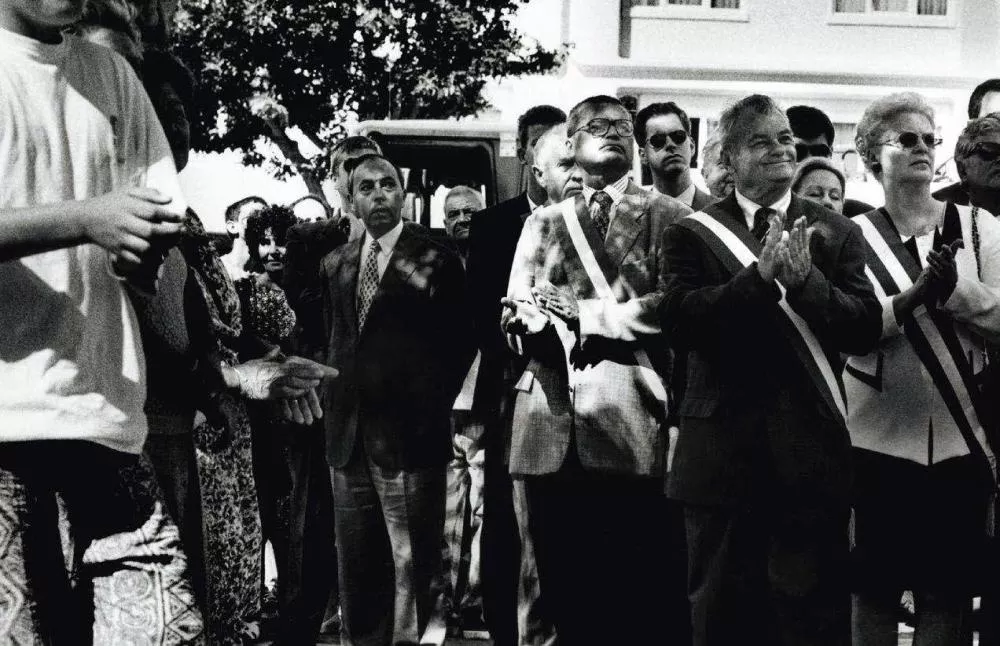 Un groupe de personnes, dont certaines portent des écharpes, semble assister à un événement en plein air. Certaines personnes applaudissent.