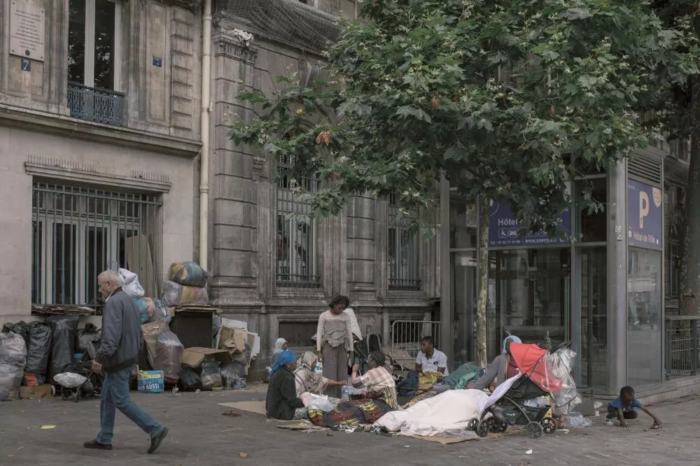 Un groupe de personnes est assis sur le trottoir devant un bâtiment, entouré de sacs et de couvertures. Un homme passe à côté, et un enfant joue près d'une poussette. Des arbres et un panneau indiquant "Hôtel de Ville" sont visibles.