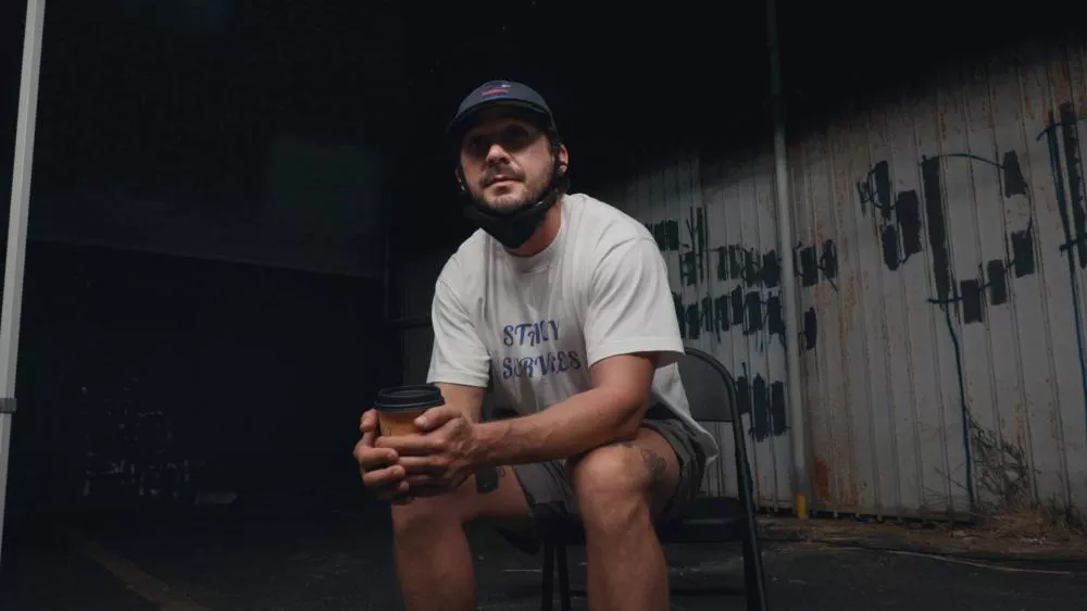 Un homme est assis sur une chaise, portant un t-shirt blanc avec l'inscription "STAY SERVED", un short et une casquette. Il tient un gobelet en carton. L'arrière-plan est sombre avec un mur métallique graffité.