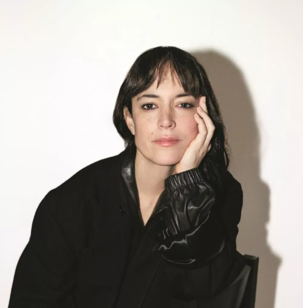 Une personne est assise sur une chaise, portant des vêtements noirs avec des détails en cuir. Elle a la tête appuyée sur sa main, avec un regard pensif. L'arrière-plan est blanc et minimaliste.