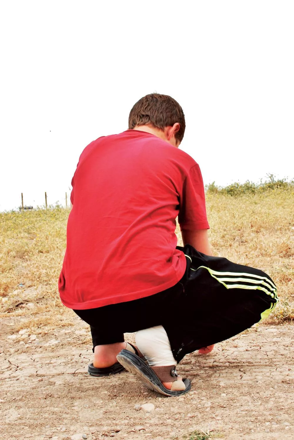 Un enfant portant un t-shirt rouge et un pantalon noir est accroupi dans un champ herbeux.