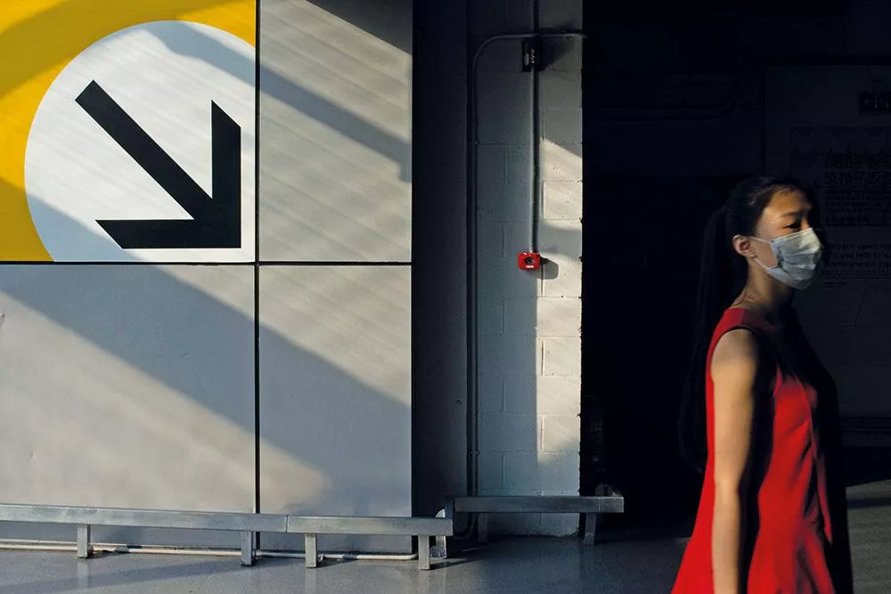 Une femme portant un masque facial marche devant un mur avec une grande flèche noire pointant vers le bas à gauche sur un fond jaune et blanc.