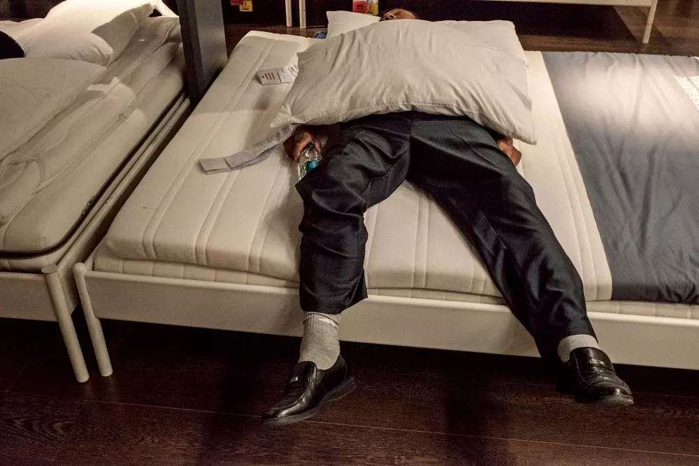 Un homme en costume est allongé sur un lit, le visage caché par un oreiller, tenant une bouteille d'eau. Ses jambes pendent du lit, et il semble être dans un environnement de magasin.