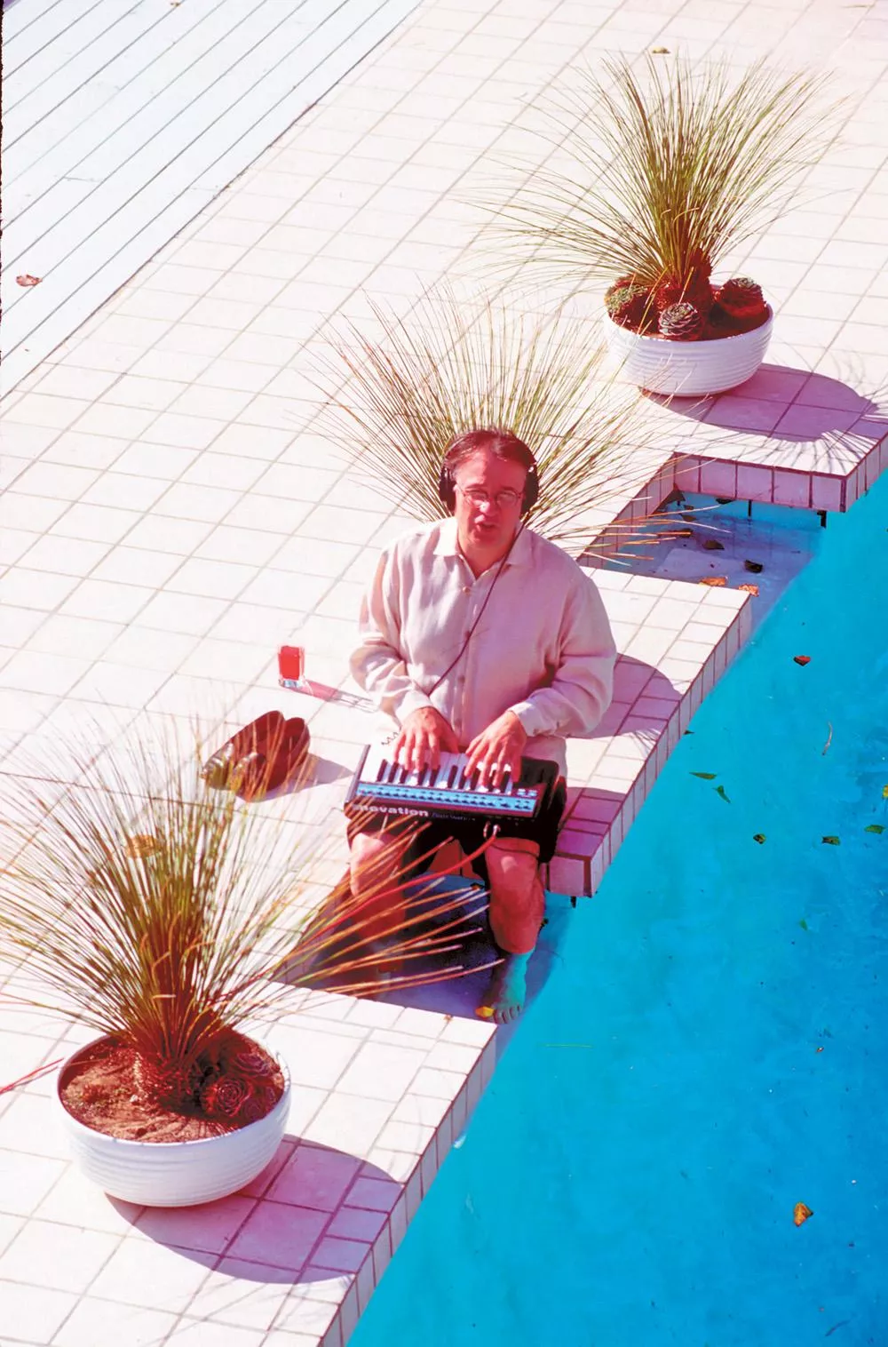 Un homme est assis près d'une piscine, jouant d'un petit clavier électronique. Il y a des plantes en pot autour de lui et un verre à côté.