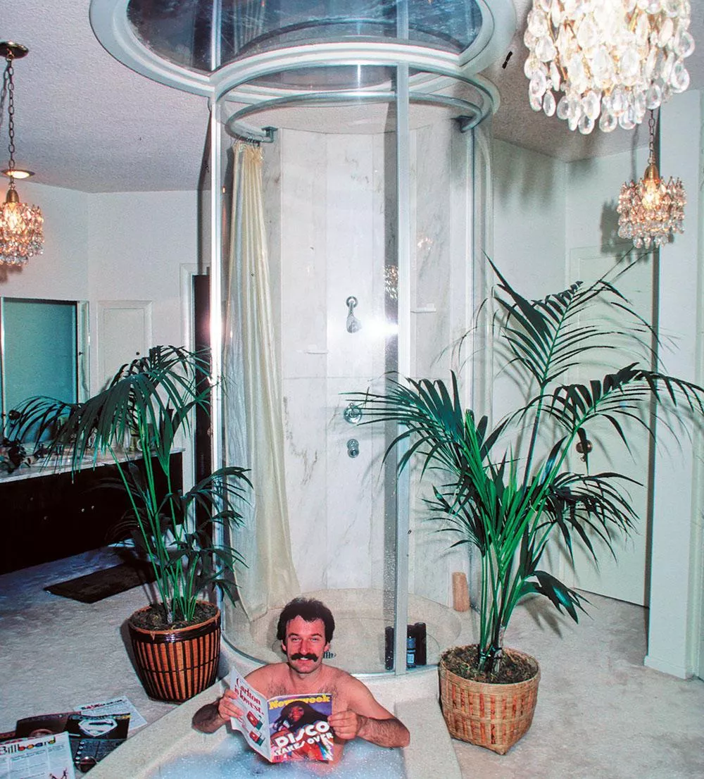 Un homme est dans une baignoire entourée de plantes, lisant un magazine intitulé "Newsweek" avec une couverture sur la musique disco. La salle de bain est décorée de lustres et d'une douche circulaire en verre.