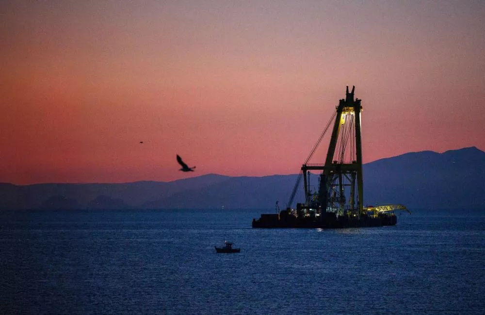 Une scène maritime au crépuscule avec une grande grue sur une plateforme en mer, un bateau plus petit à proximité, et un oiseau en vol. L'horizon montre des montagnes sous un ciel aux teintes orangées et bleutées.