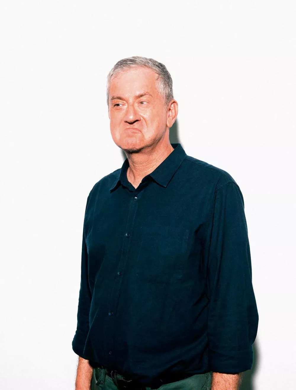 Un homme aux cheveux gris, portant une chemise bleue, fait une grimace avec les lèvres pincées. Il se tient debout devant un fond blanc.