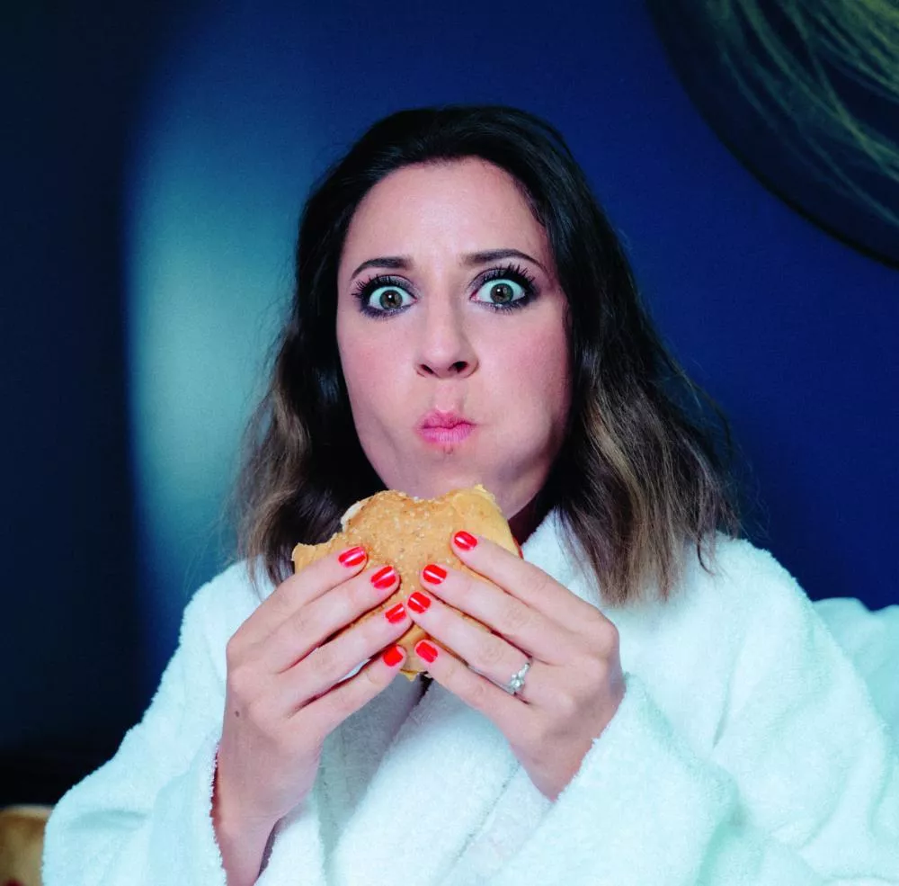 Une personne en peignoir blanc mange un sandwich avec une expression surprise.