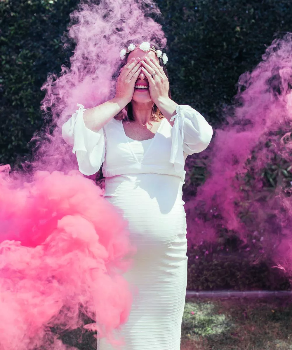 Une femme en robe blanche, enceinte, se tient dans une nuée de fumée rose, les mains couvrant son visage.