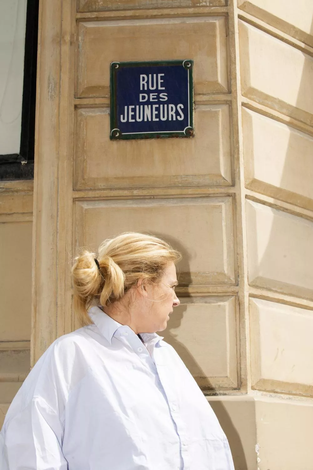 Une femme se tient devant un mur beige avec une plaque indiquant "Rue des Jeûneurs".
