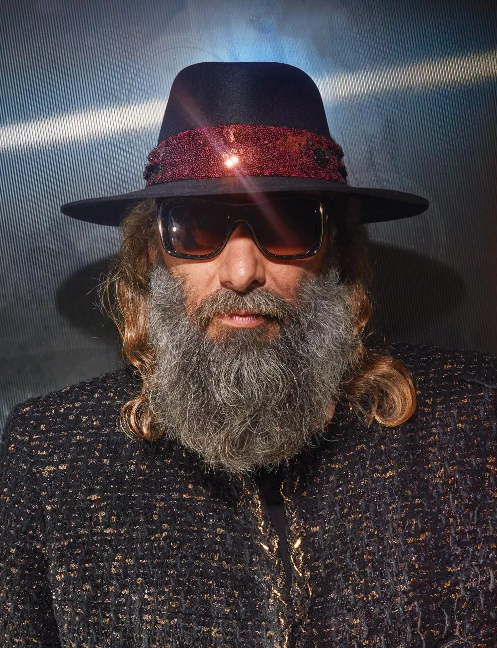 Un homme avec une barbe grise et des cheveux longs porte un chapeau noir orné d'un ruban rouge brillant et des lunettes de soleil. Il est habillé d'une veste texturée sombre.
