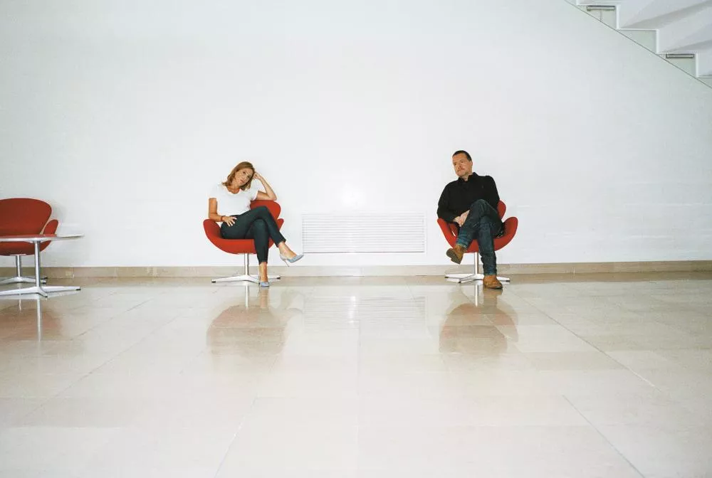 Deux personnes sont assises sur des fauteuils rouges dans une pièce aux murs blancs. Elles sont éloignées l'une de l'autre, avec un espace vide entre elles.