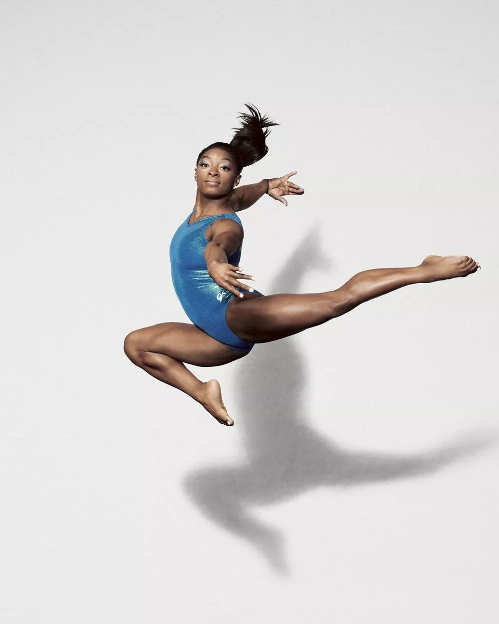Une gymnaste en justaucorps bleu est en plein saut, exécutant une pose élégante et dynamique avec les bras et les jambes étendus.