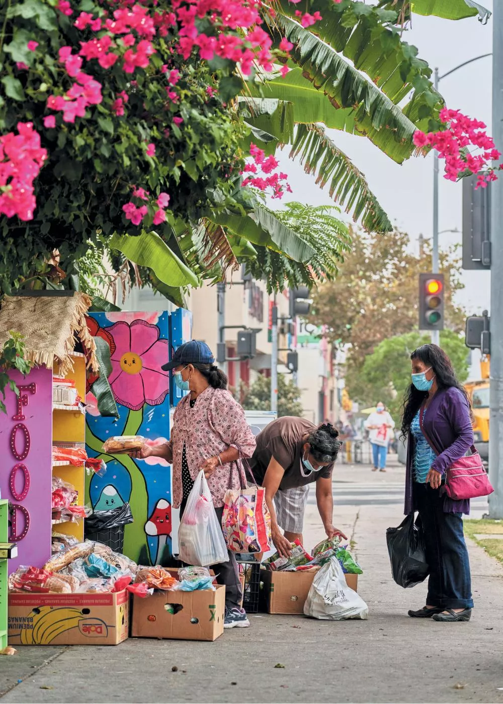 Trois personnes portant des masques faciaux sont en train de choisir des aliments dans une étagère colorée en bord de rue, entourée de fleurs roses et de feuillage.