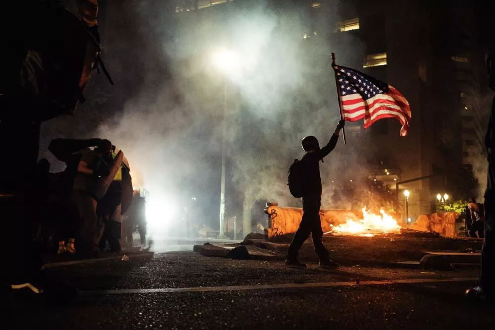 Une personne tient un drapeau américain lors d'une manifestation nocturne, avec des feux et de la fumée en arrière-plan.