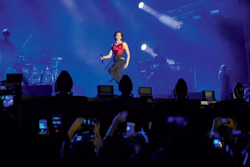Une artiste en concert sur scène, tenant un micro, avec des musiciens en arrière-plan et un public prenant des photos.
