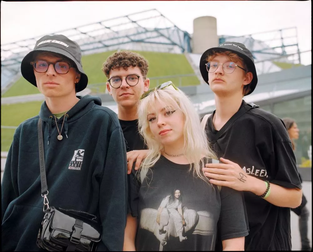 Un groupe de quatre jeunes, trois hommes et une femme, posant ensemble. Ils portent des vêtements décontractés et des lunettes, et deux d'entre eux portent des chapeaux bob. L'arrière-plan montre une structure architecturale moderne.