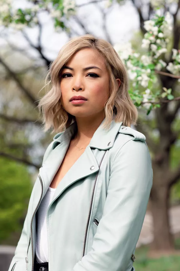 Une personne aux cheveux blonds ondulés porte une veste en cuir vert clair. L'arrière-plan montre des arbres en fleurs.