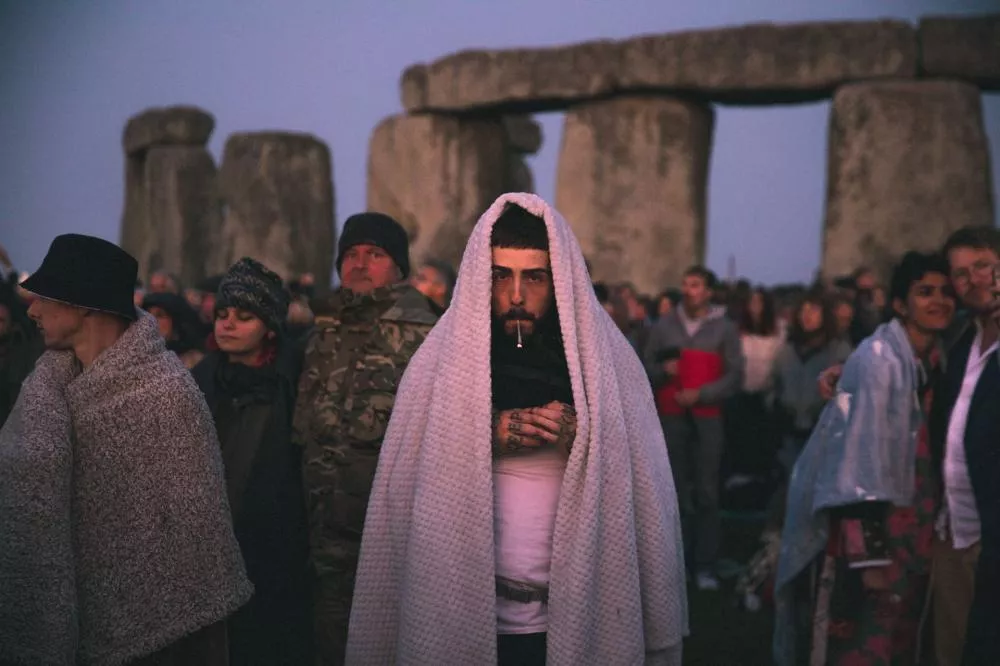 Un groupe de personnes est rassemblé devant Stonehenge. Une personne au centre est enveloppée dans une couverture blanche, tenant un petit objet dans ses mains. L'ambiance semble être celle d'un rassemblement ou d'un événement.