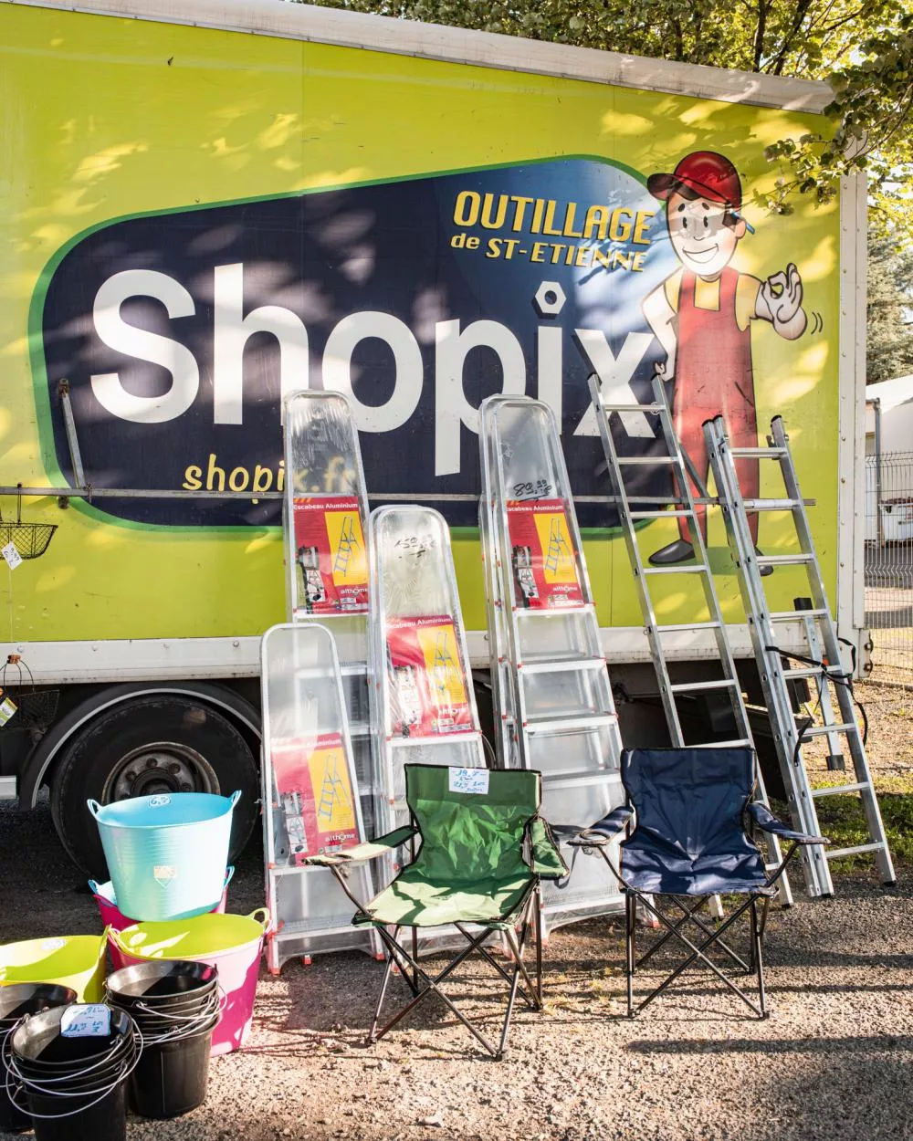 Un camion avec l'inscription "Shopix" et "Outillage de St-Étienne" est garé. Devant, il y a des échelles, des seaux colorés, et des chaises pliantes exposées.