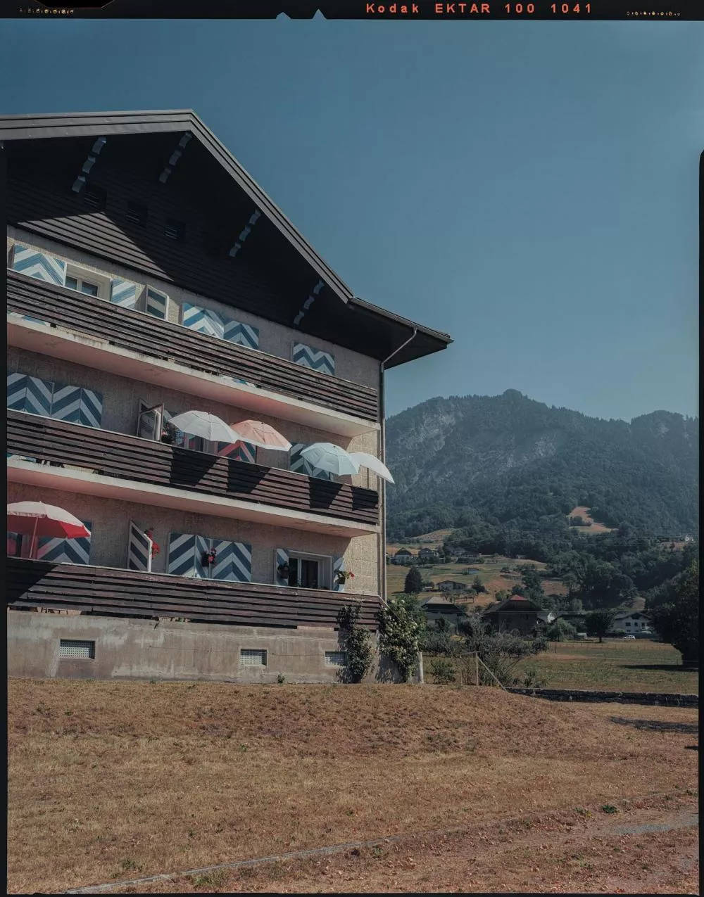 Un bâtiment résidentiel avec des balcons ornés de parasols colorés, situé dans un paysage montagnard avec des collines verdoyantes à l'arrière-plan.