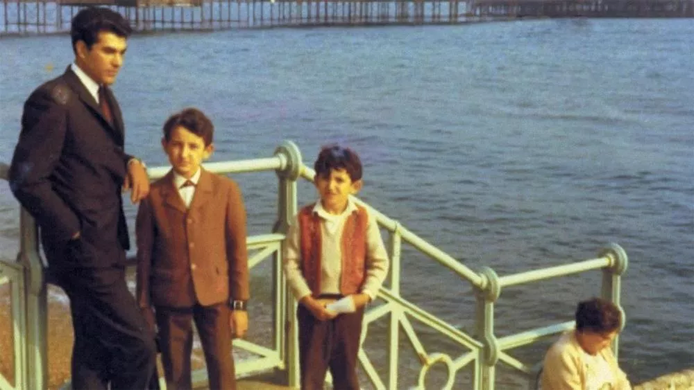 Un homme et deux enfants posent près d'une balustrade au bord de l'eau, avec une personne assise en train de lire ou écrire. En arrière-plan, on aperçoit une jetée.