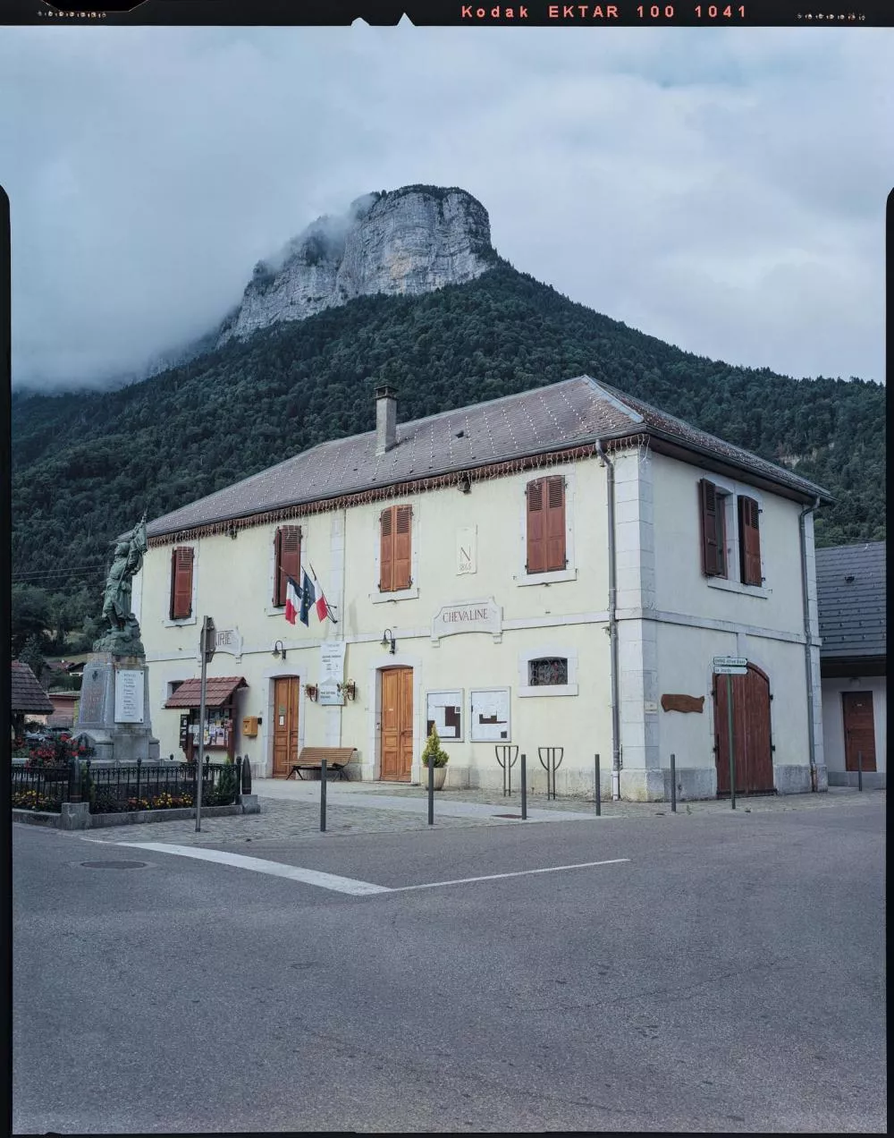 Un bâtiment avec des volets rouges et un toit en tuiles, situé devant une montagne couverte de végétation. Il y a des drapeaux accrochés à l'avant du bâtiment et une statue sur la gauche. Le ciel est nuageux.