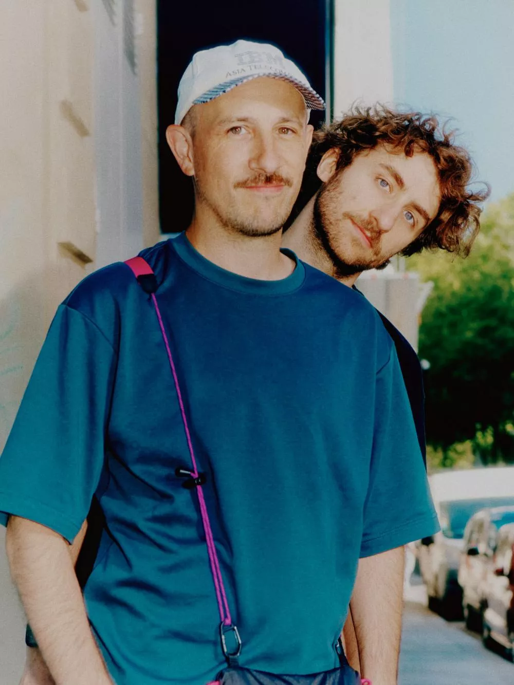 Deux hommes posent ensemble, l'un portant une casquette blanche et un t-shirt bleu avec une sangle rose. Ils semblent être dehors, avec des arbres en arrière-plan.