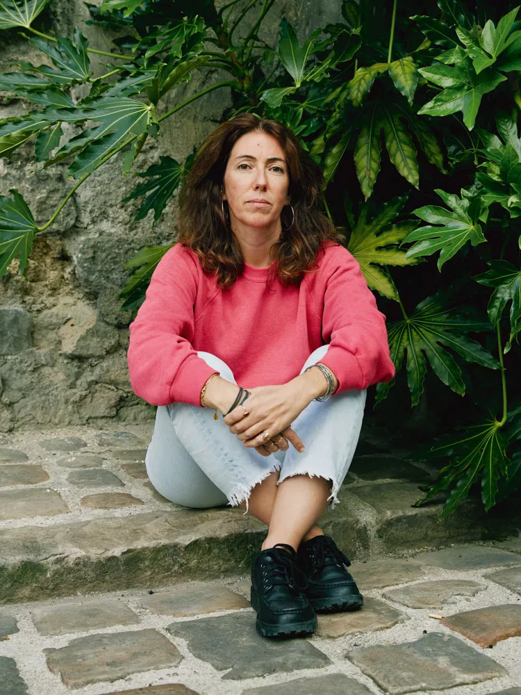 Une personne est assise sur des marches en pierre, entourée de feuillage dense. Elle porte un pull rose, un jean clair et des chaussures noires.