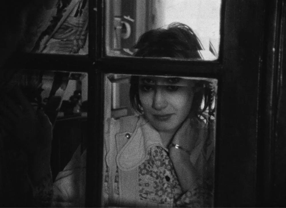 Une femme regarde à travers une fenêtre, avec un air pensif et légèrement souriant. Elle porte une chemise à motifs floraux et un bracelet. L'image est en noir et blanc.