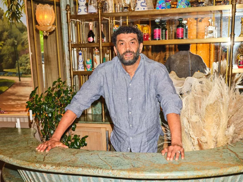 Un homme se tient derrière un comptoir en marbre vert, entouré de décorations et d'étagères contenant des bouteilles et divers objets colorés. L'arrière-plan montre un décor intérieur avec des plantes et un mur orné.