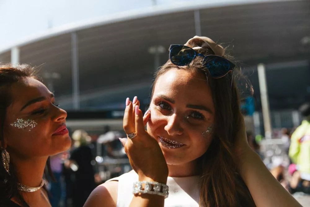 Deux personnes souriantes avec des paillettes sur le visage, l'une portant des lunettes de soleil sur la tête. L'arrière-plan montre une structure moderne et floue, suggérant un événement en extérieur.