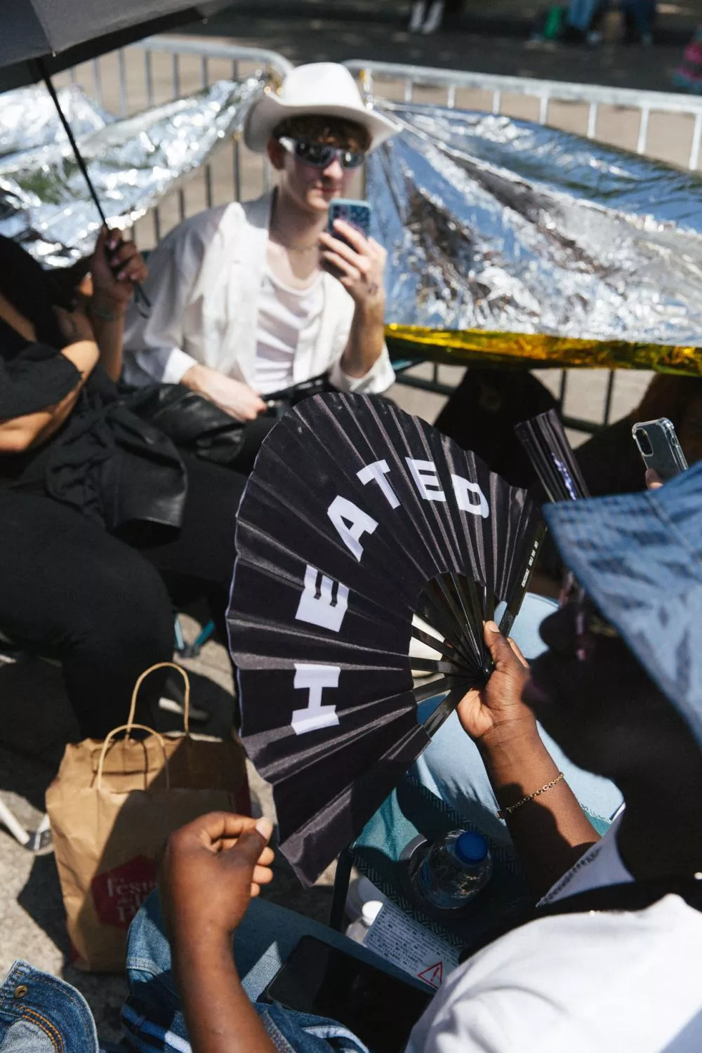 Des personnes assises à l'extérieur, certaines utilisant des parapluies et des éventails pour se protéger du soleil. Une personne tient un éventail avec le mot "HEATED". Une autre personne est habillée en blanc avec un chapeau et des lunettes de soleil, regardant son téléphone.