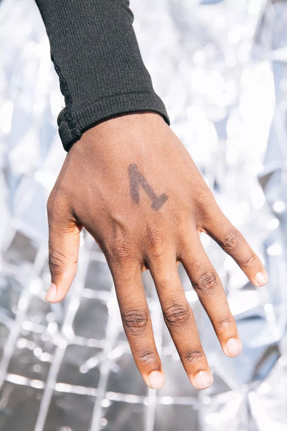 Une main avec un chiffre "1" dessiné dessus, portant un vêtement noir à manches longues. L'arrière-plan est flou et lumineux.