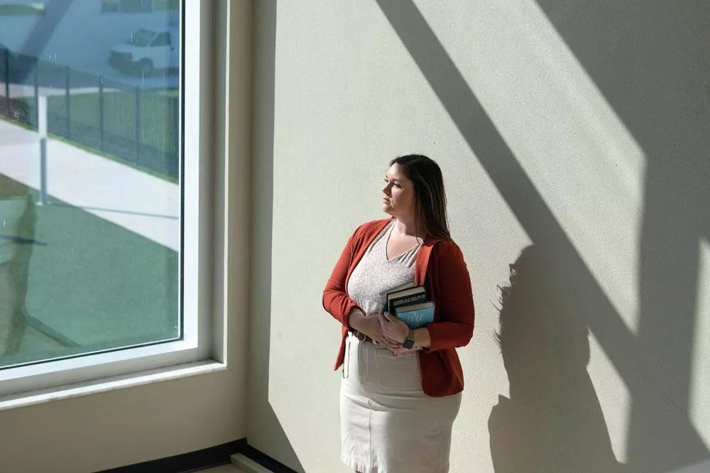 Une personne se tient près d'une fenêtre, tenant des livres. Elle porte un cardigan rouge et une jupe beige. La lumière crée des ombres sur le mur.
