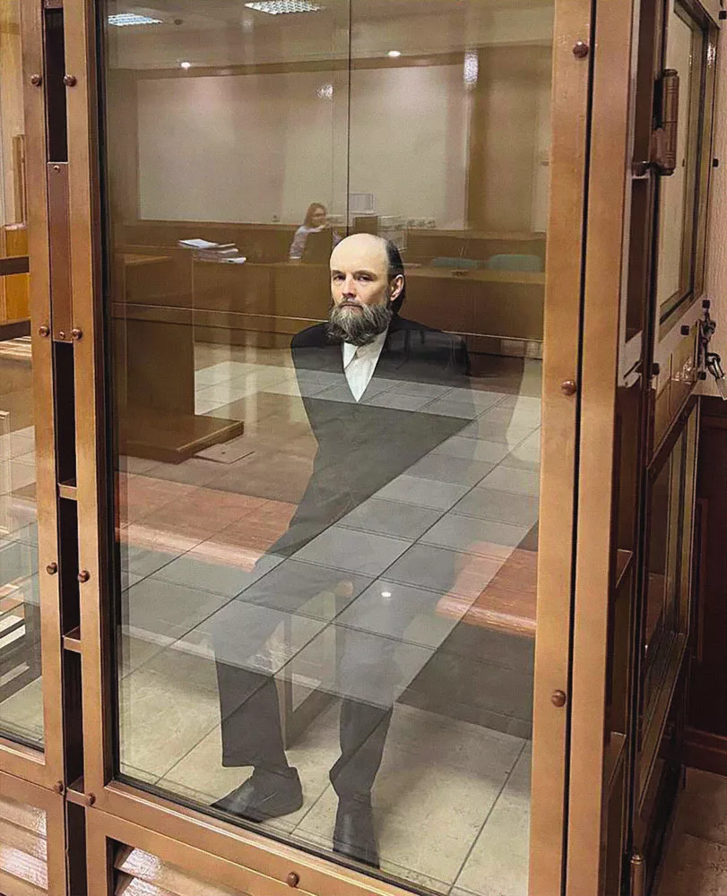 Un homme barbu en costume est assis dans un box en verre dans une salle d'audience.