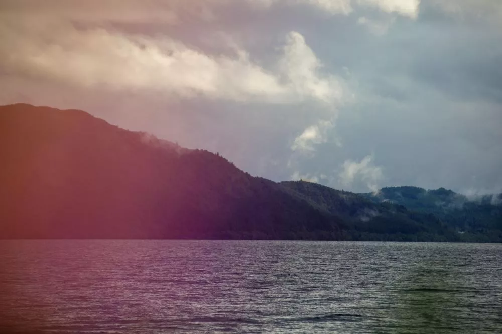 Un paysage de lac avec des collines boisées à l'arrière-plan sous un ciel nuageux, avec une teinte rosée sur la gauche de l'image.