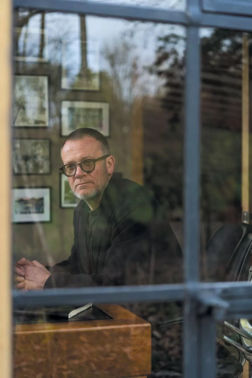 Un homme avec des lunettes est assis à l'intérieur, regardant par la fenêtre. Des cadres sont accrochés au mur derrière lui.