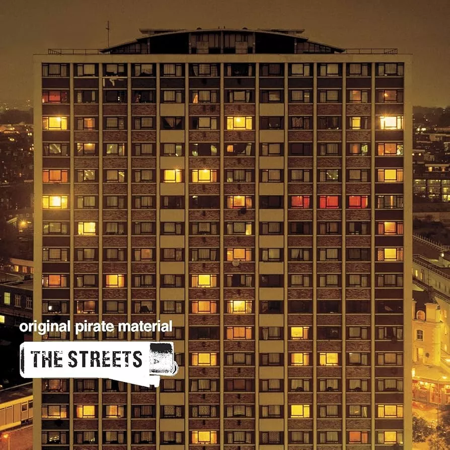 Un immeuble résidentiel éclairé la nuit, avec des fenêtres illuminées. Le texte "original pirate material" et "THE STREETS" est visible en bas à gauche.