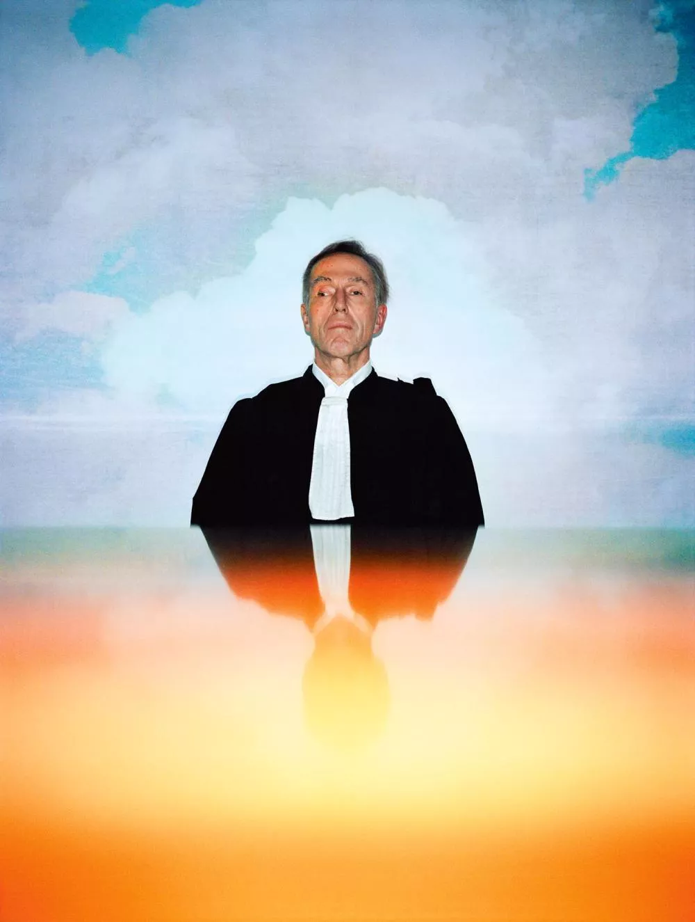 Un homme en tenue formelle se tient devant un fond de ciel nuageux, avec un reflet coloré en bas de l'image.